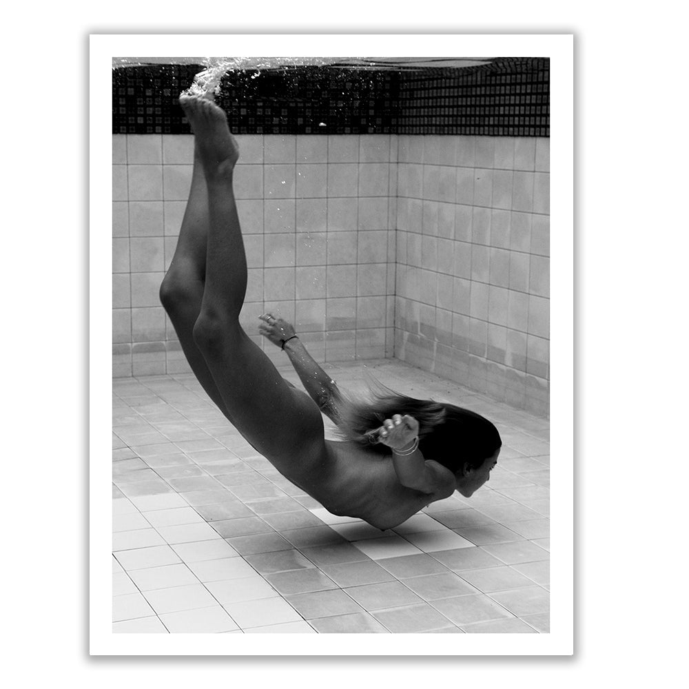 A person stands against a brick wall, holding a large framed black-and-white photograph. The photo shows a person floating upside down in water. The holder's face is obscured by the frame.