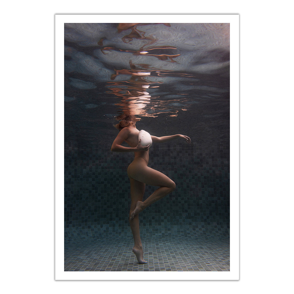 A person in a swimsuit poses gracefully underwater, standing on one leg with the other bent, reaching toward the water's surface, creating gentle ripples and reflections above.