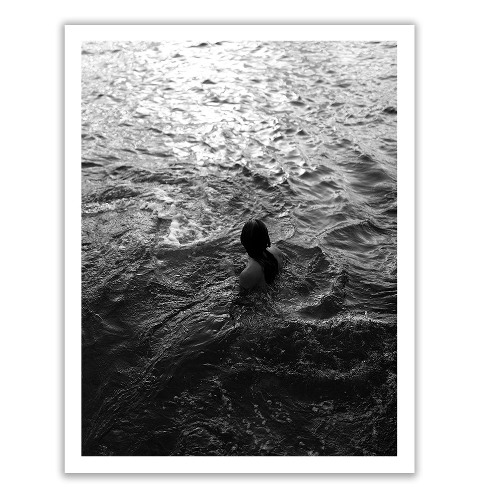 Sunrise Swim #11 by Francesca Owen captures a black and white photo of a person in the ocean viewed from behind. The individual stands waist-deep in the water, surrounded by rippling waves with light reflecting on the surface. Printed on fine art paper using archival pigments, this piece includes a certificate of authenticity.