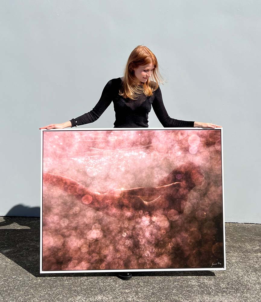 A person with long, red hair is wearing a black outfit and holding a large, rectangular piece of art titled "Blush 02" by Francesca Owen. The artwork, printed on fine art paper using archival pigments, features an abstract design with pink and brown hues creating a textured, blurry effect against a plain, light-colored wall.