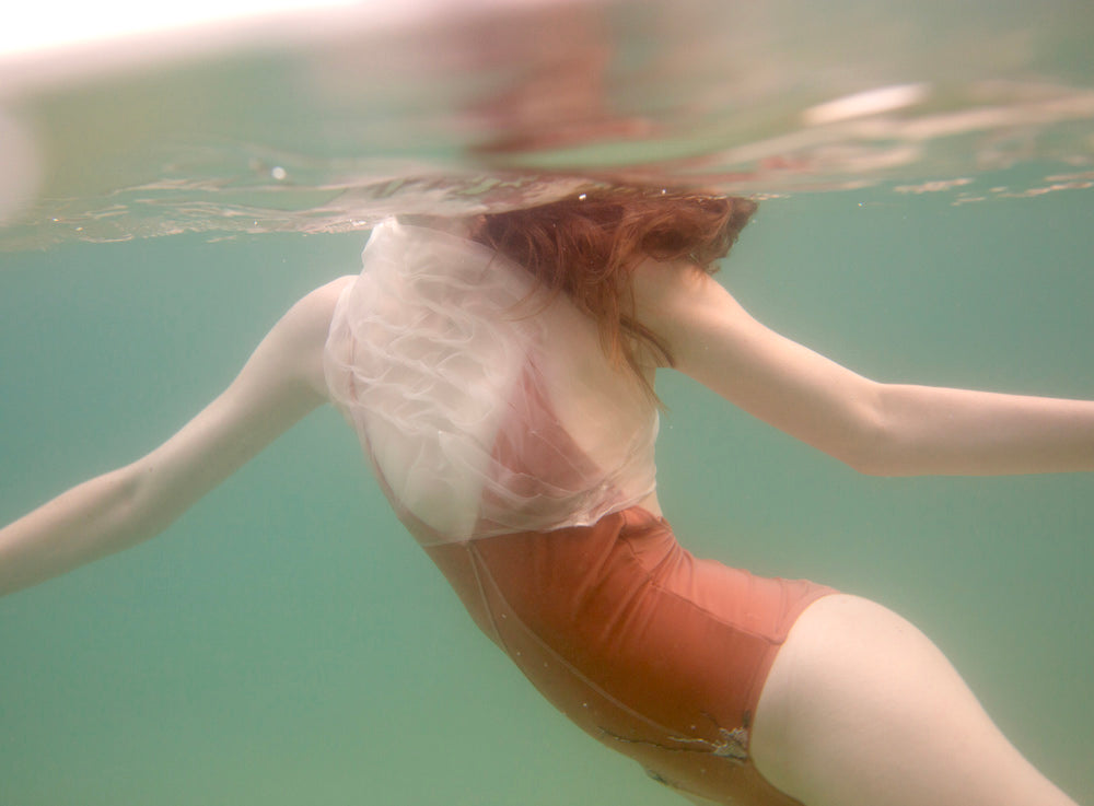 A person elegantly swimming underwater, wearing Francesca Owen's Feminine swimsuit in a rust hue with sheer, ruffled fabric, as their long hair streams behind them. The transparent green water and gentle ripples create an ethereal feeling of motion akin to art preserved on fine art paper.