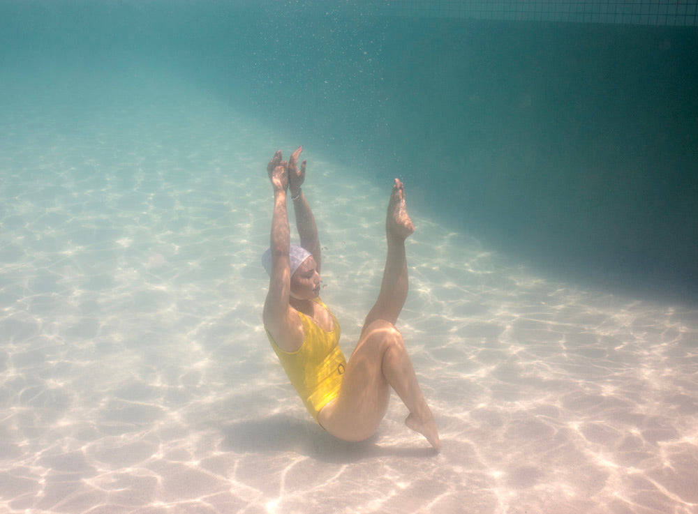 A person in a yellow swimsuit performs a graceful gymnastics move underwater in a pool, with arms extended upwards, knees slightly bent, and feet pointing forward, creating an elegant floating pose. The water is crystal clear and sunlight filters through, casting intricate patterns on the pool floor. This serene moment is exquisitely captured in "Halcyon Daze 3" by Francesca Owen, ideal for fine art prints.