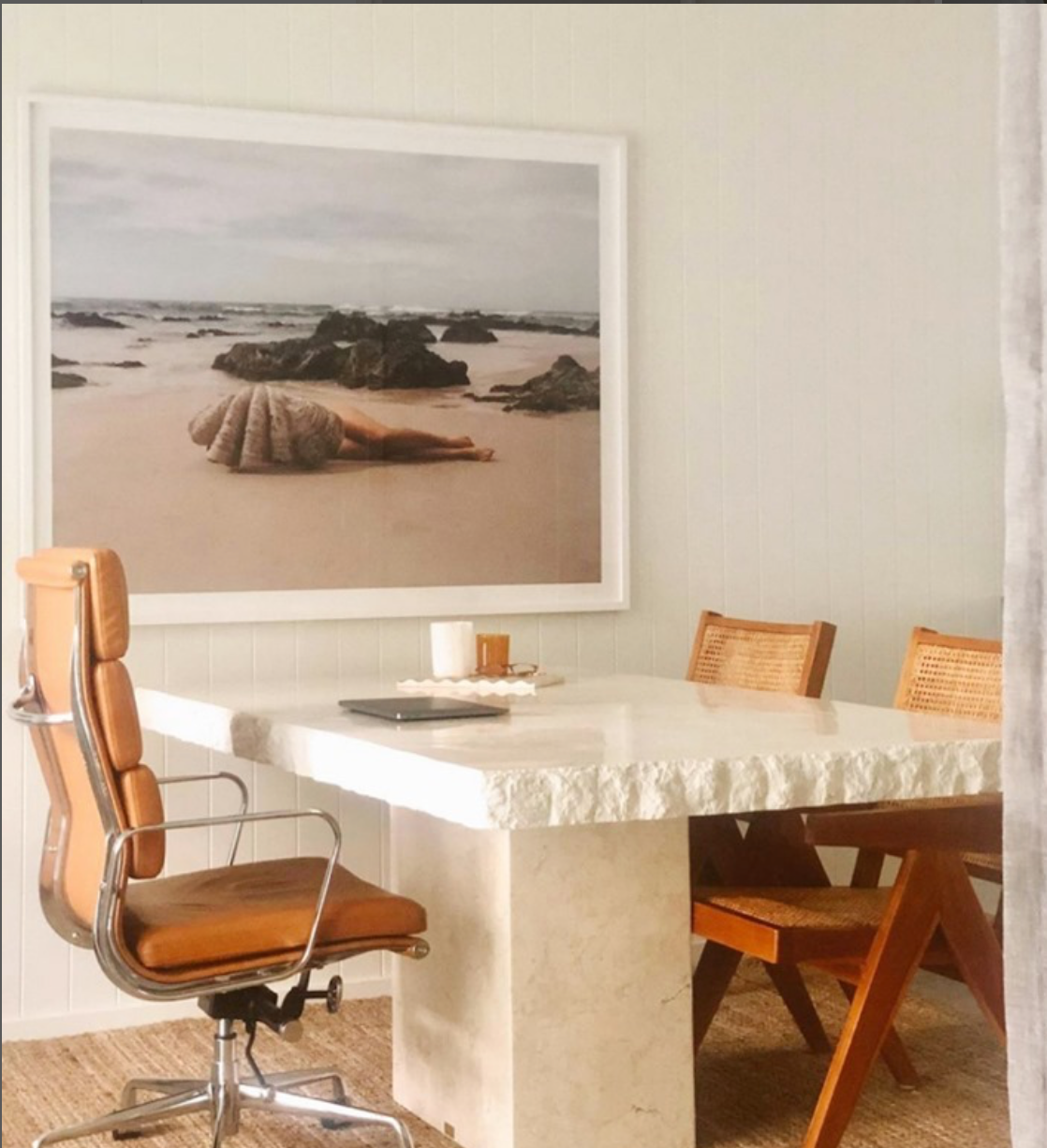 A minimalist dining area features a rectangular stone table with two wooden chairs and a leather swivel chair. Dominating the space is "The Beauty Within" by Francesca Owen, a large framed photograph printed on fine art paper using archival pigments, depicting a sandy beach with a large seashell and rocks. The room exudes a coastal, serene atmosphere.
