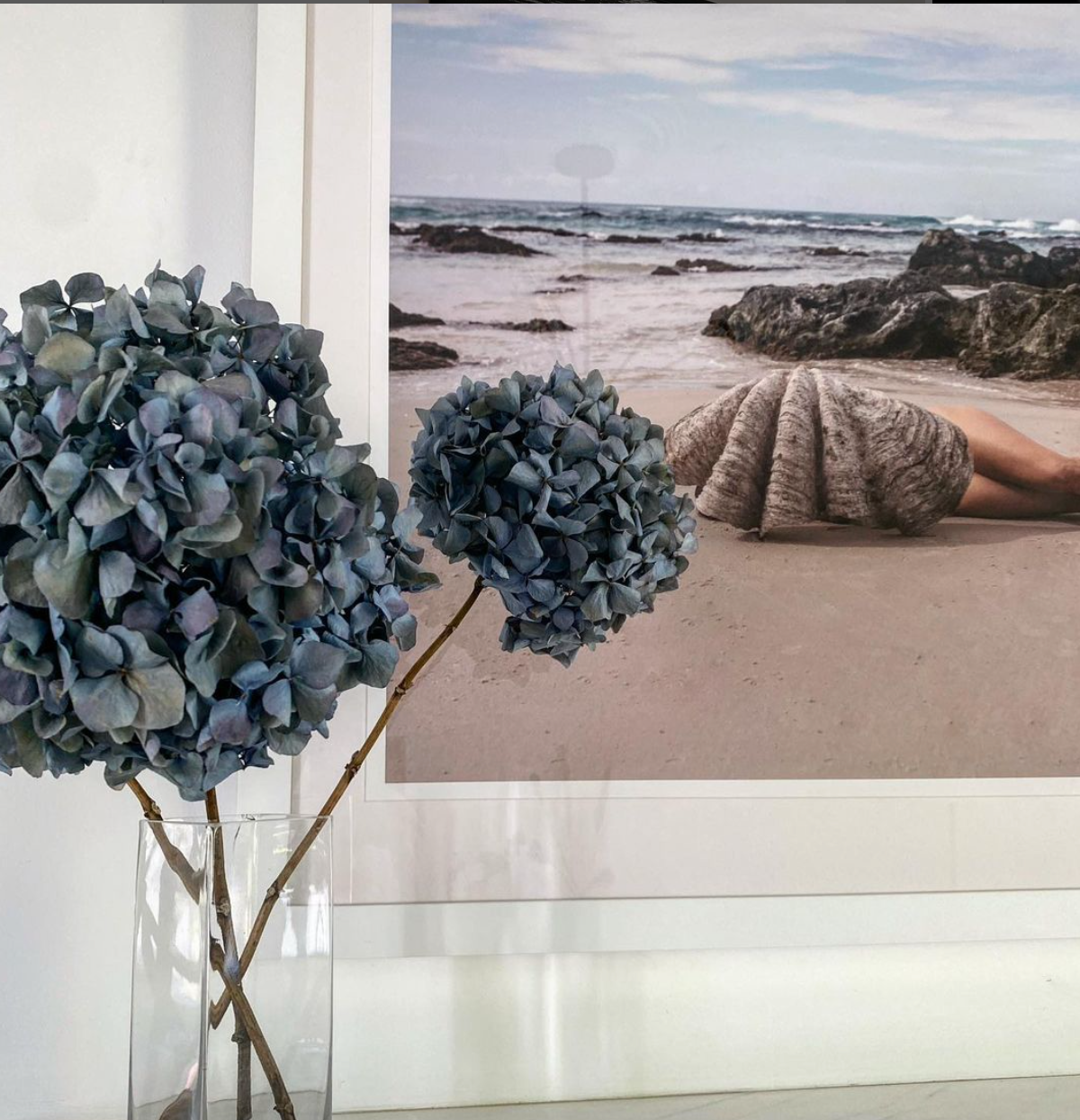 A framed photograph titled "The Beauty Within" by Francesca Owen, depicting a beach with rocks and a person wrapped in a large seashell, leans against a wall. In the foreground, there is a glass vase holding two branches of dried blue hydrangeas. Printed with archival pigments on fine art paper, this piece promises durability and vibrancy for years to come.