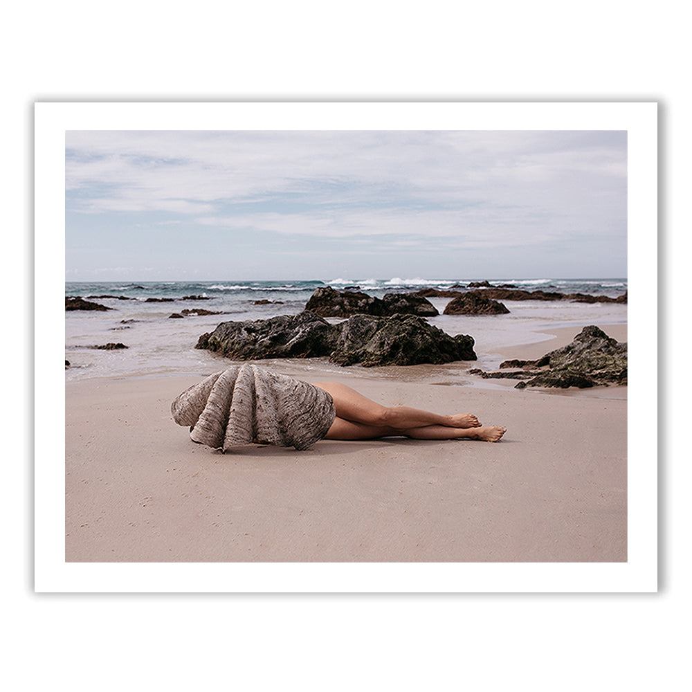 A person lies on a sandy beach with their legs exposed, the rest of their body covered by a large seashell. Around them, several rocks are scattered along the shoreline, and the ocean waves are visible in the background under a partly cloudy sky. This scene would make for stunning fine art paper prints using archival pigments from Francesca Owen's "The Beauty Within.