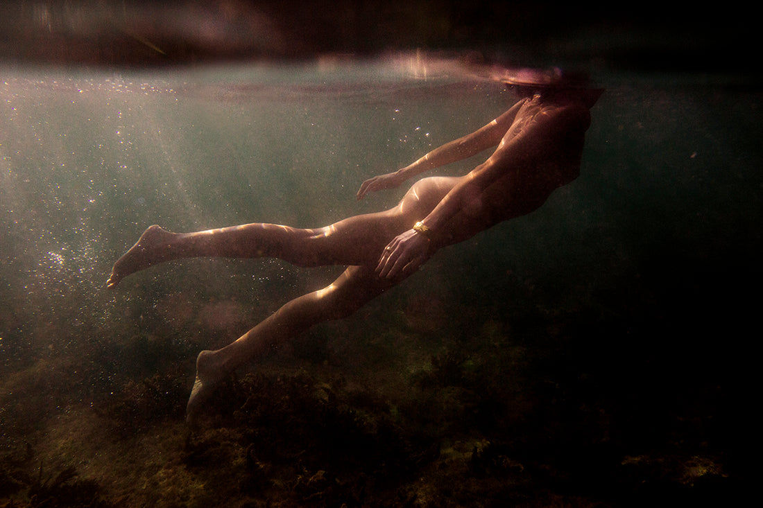 A person swimming underwater in a slightly upward direction in a dimly lit aquatic environment. The water appears dark with sunlight filtering through the surface, illuminating the swimmer's body and the surrounding area, capturing an ethereal moment worthy of archival pigments on fine art paper. This scene evokes the same delicate allure as Blush 03 by Francesca Owen.
