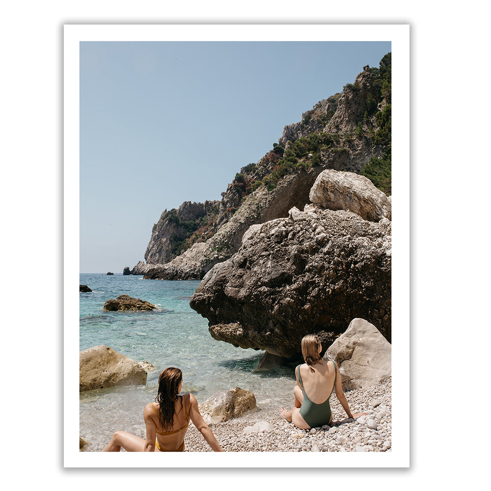 Two women sit on a rocky shoreline facing the sea, one in a yellow bikini and the other in a green one-piece swimsuit. Large rocks surround the clear blue water, with a steep, rocky hill covered in greenery rising in the background under a clear blue sky. This scene is captured beautifully in "Capri Mermaids" by Francesca Owen, available in custom sizes with archival pigments and accompanied by a certificate of authenticity.