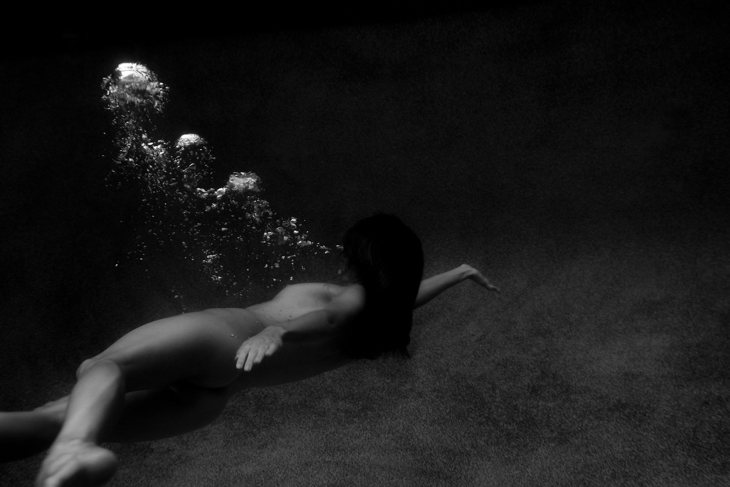 The monochromatic image, "Breath Out" by Francesca Owen, depicts a person with long hair submerged underwater, surrounded by bubbles ascending towards the surface. Printed on fine art paper, the ambient lighting and smooth gradients create a serene and ethereal atmosphere. This artwork is available in custom sizes and framing options.