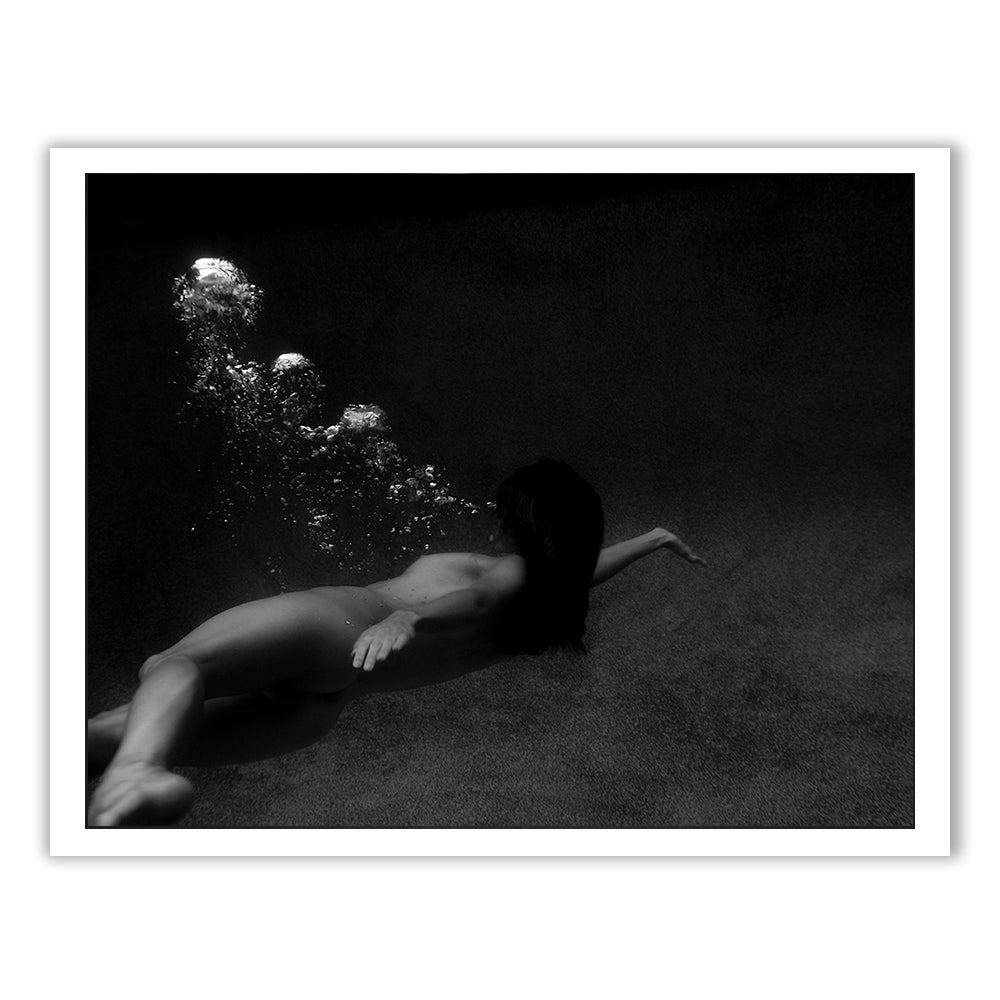 Breath Out by Francesca Owen captures a serene and ethereal moment in a black-and-white photograph. The image shows a person swimming underwater with arms extended forward, surrounded by dark, shadowy water and trailing bubbles. This timeless piece is printed on fine art paper for an exquisite finish.
