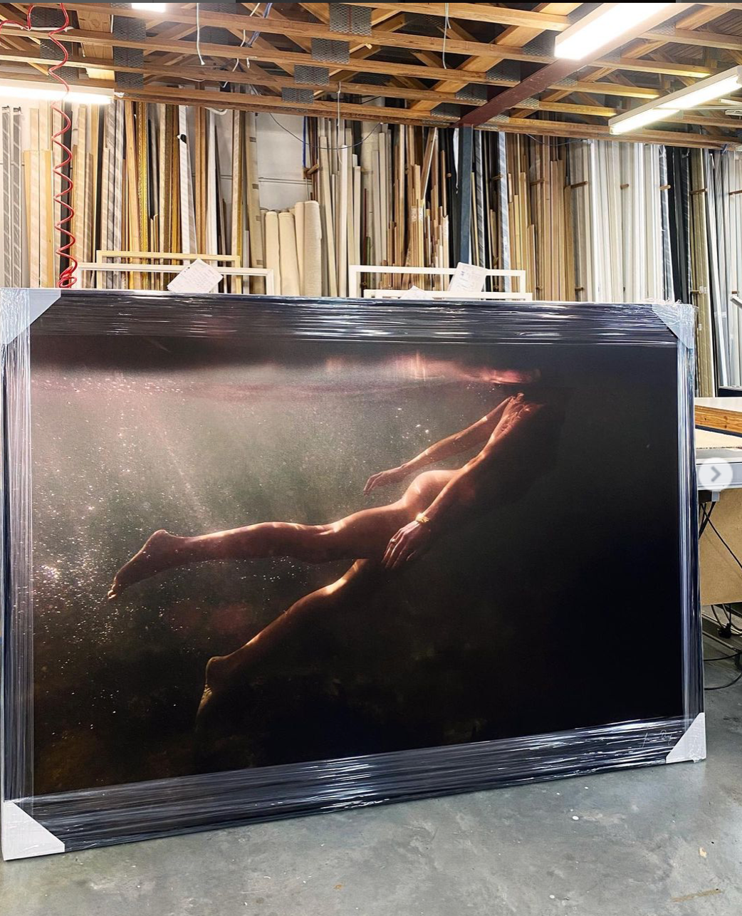 A large, framed fine art print titled *Blush 03* by Francesca Owen features a person submerged underwater, partially illuminated by light. The print leans against a wall in a workshop or studio space, depicting the person floating horizontally with archival pigments creating a dramatic, ethereal effect on their form.