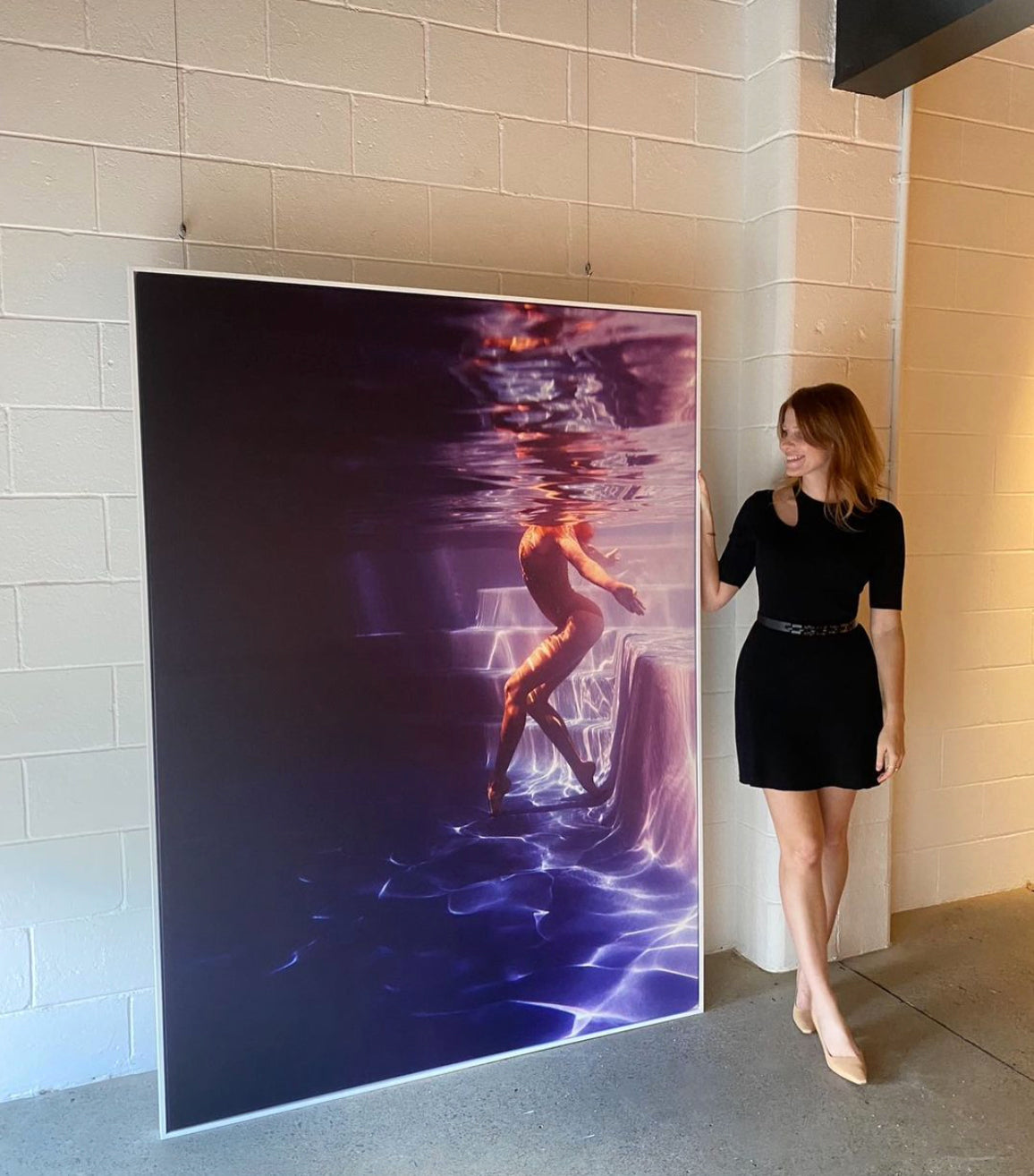 A woman in a black dress stands next to a large, framed artwork hanging on a white brick wall. The fine art print, named Glisten 06 by Francesca Owen, depicts a person underwater surrounded by shimmering light, creating a dreamy, ethereal effect. Smiling, she admires the piece's custom framing that perfectly complements the scene.