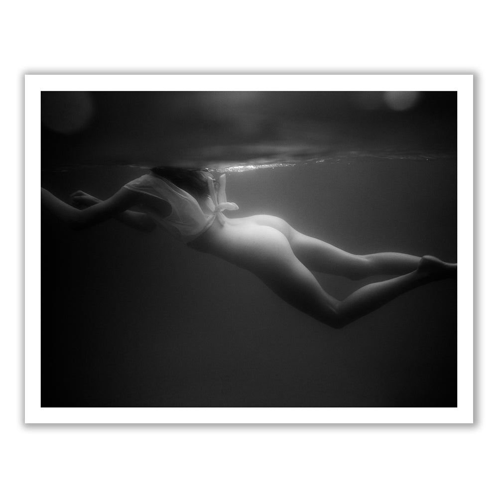 Gliding #1 by Francesca Owen is a black and white photograph capturing a person swimming underwater in a dramatic scene. The swimmer is positioned horizontally, facing downwards and slightly to the left, with their body partially immersed in water. Light and shadows play across the image, adding to its striking effect. This fine art print comes with a certificate of authenticity.