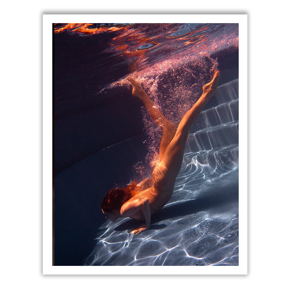Image of a person underwater in a swimming pool, diving headfirst towards the pool steps. Air bubbles rise from their body as they descend, and the water reflects light, creating patterns. The person is surrounded by the deep blue hue of the water. Available in custom sizes and framing with a certificate of authenticity. This piece is titled "Glisten 03" by Francesca Owen.