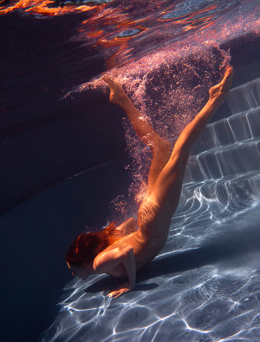 Glisten 03 by Francesca Owen captures the serene moment of a person diving toward the bottom of a pool, with water shimmering over their body and air bubbles trailing behind them. The light from above casts a warm glow, enhancing the fluid motion. This artwork is available on fine art paper with custom sizes and framing options.