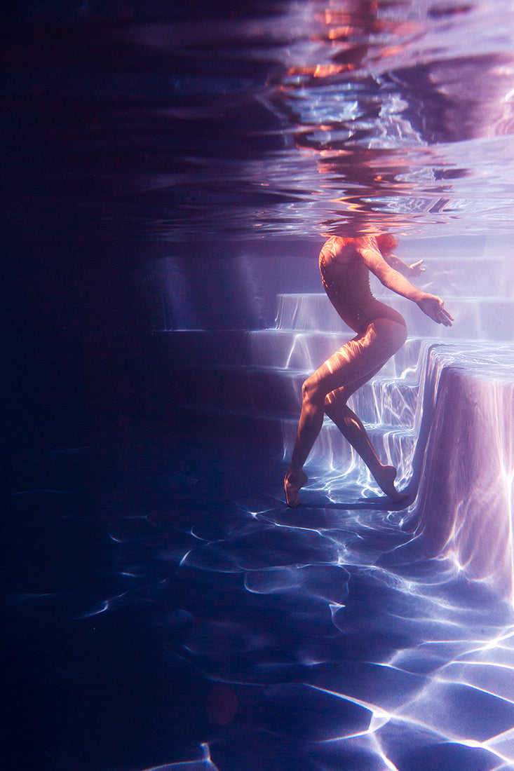 Underwater view of a person in a swimming pool, illuminated by the mesmerizing colors of Francesca Owen's Glisten 06. The person is gracefully posing with one knee bent, touching the steps, while their upper body reaches towards the water's surface, distorted by reflections that shimmer like archival pigments on fine art paper.
