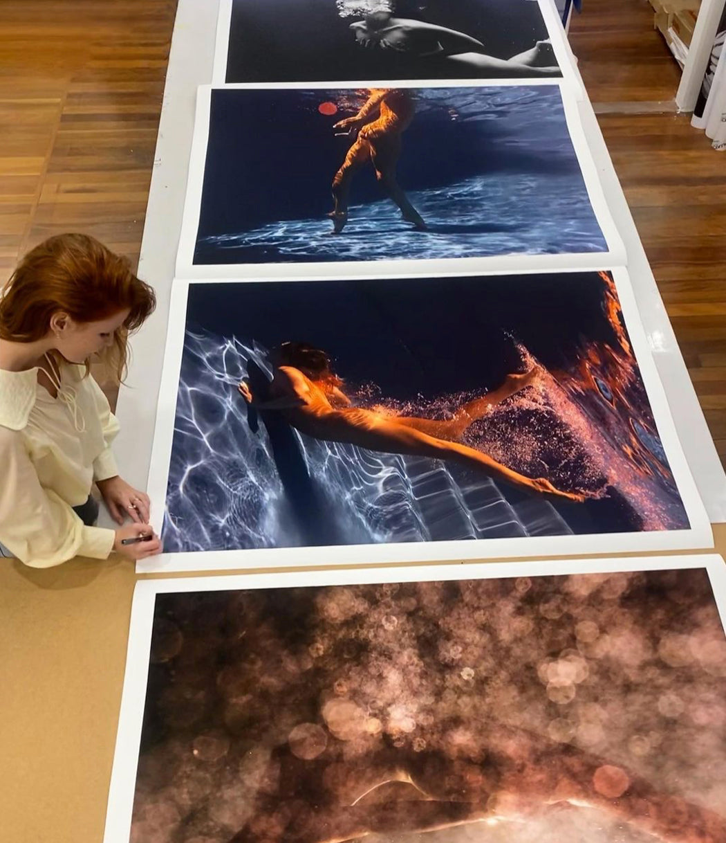 A woman with long red hair is seen inspecting and signing large prints of "Glisten 03," an underwater photograph by the artist Francesca Owen, showcased on fine art paper laid out on a table. Each image depicts models immersed in water, with varying lighting effects creating ethereal and dynamic scenes. She also includes a certificate of authenticity with each piece.