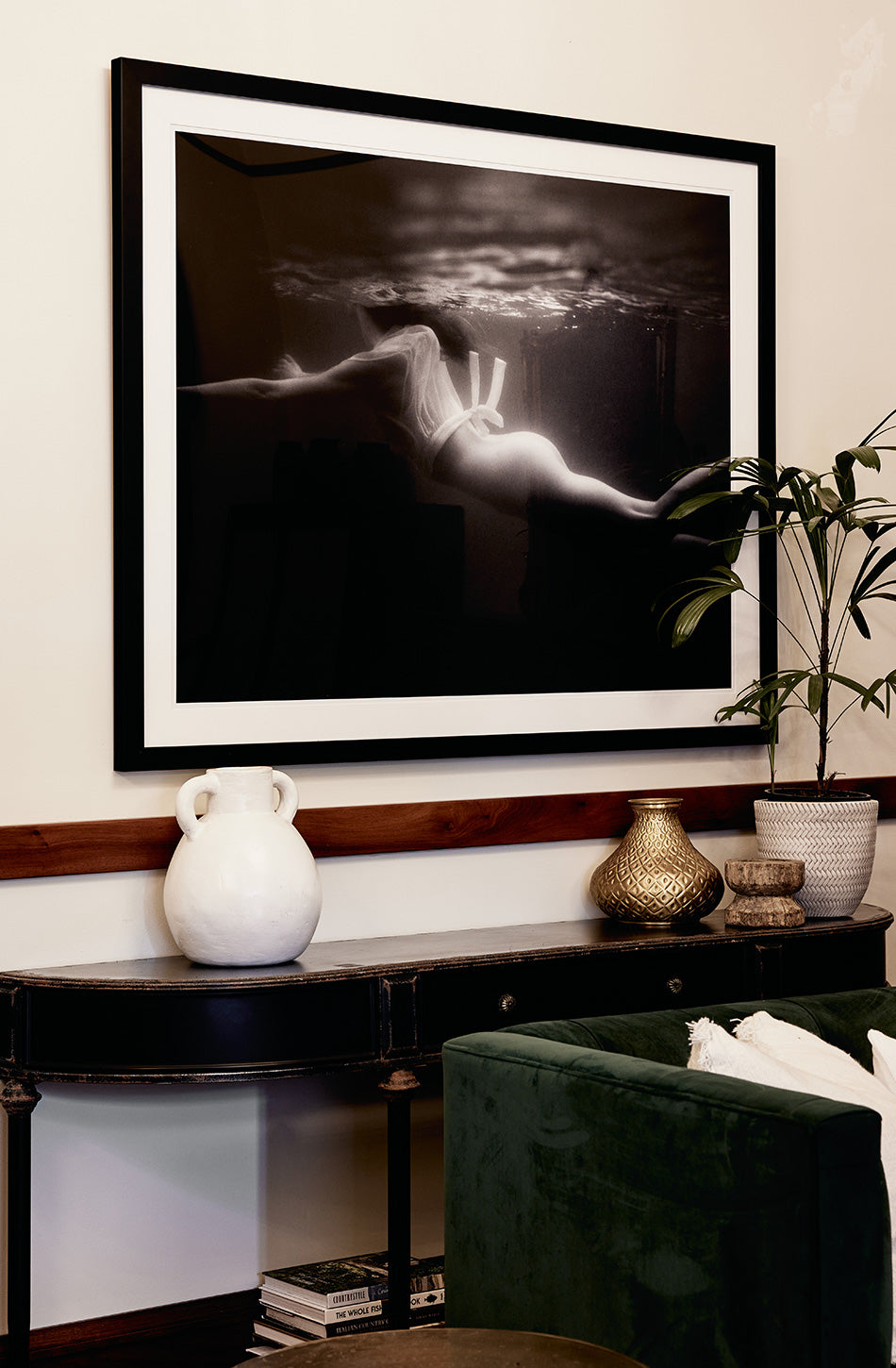 A stylish living space features a dark wooden console table adorned with a white ceramic vase, a gold decorative piece, and a potted plant. Above, Gliding #1 by Francesca Owen, a large black and white artwork depicting an underwater scene, is framed and mounted on a cream-colored wall. The artwork is printed with archival pigments for lasting beauty.