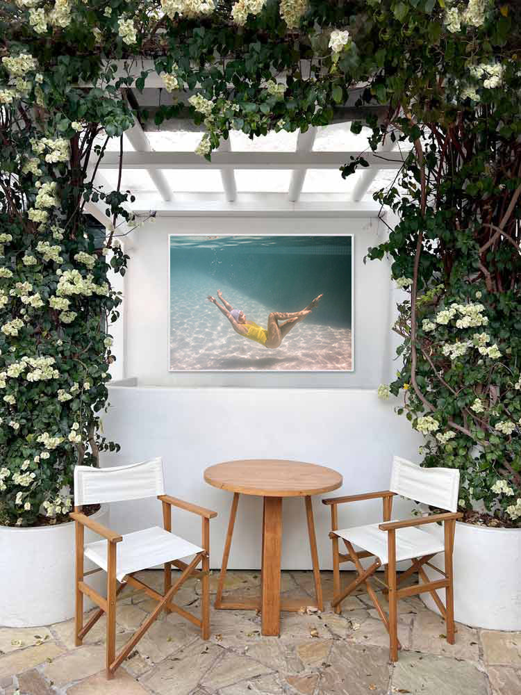 A small patio with two white chairs and a round wooden table sits under a pergola. On the white wall behind, "Halcyon Daze #1" by Francesca Owen—a striking signed photo of a person swimming underwater—is displayed.
