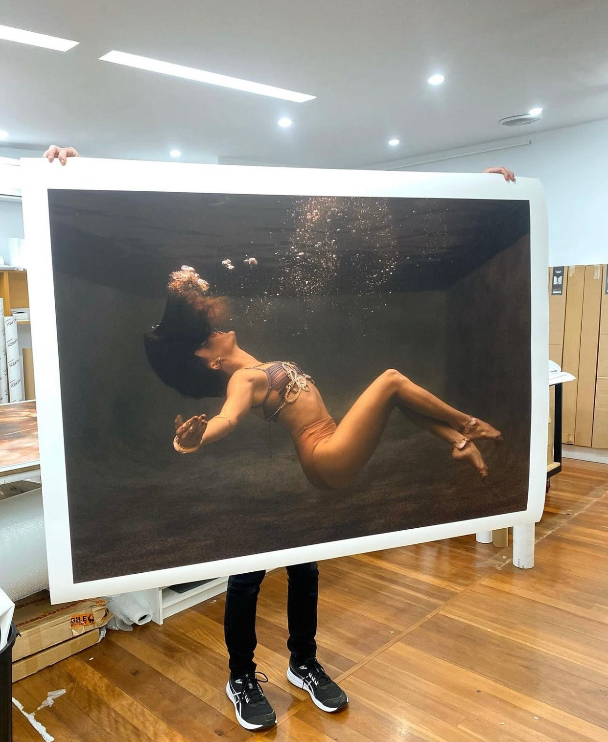 A person holds a large print of *Show Time Part 3, #3* by Francesca Owen, featuring an image of a woman floating gracefully underwater amidst bubbles. This captivating photograph is rendered on fine art paper using archival pigments and enhances the ambiance in a room with wooden floors and bright overhead lights.