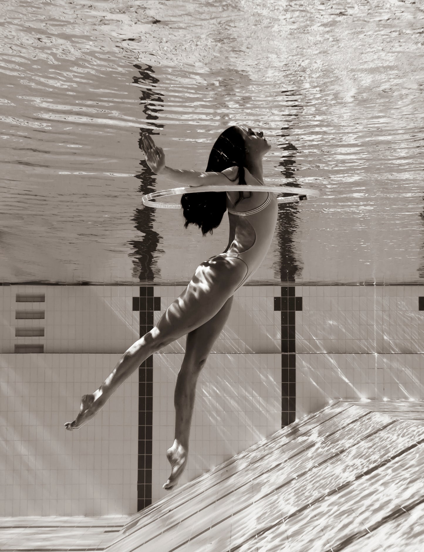 Underwater shot of a woman in a swimsuit gracefully posing with her arms extended and legs slightly bent, as if dancing in water. Her long hair flows freely, and the pool tiles and water reflections create an artistic backdrop. This scene has been captured beautifully by Francesca Owen in her work titled "Flying Solo 01," worthy of fine art prints that come complete with a certificate of authenticity.