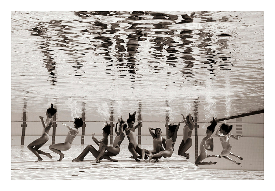 A group of people dressed in swimsuits are underwater at the edge of a swimming pool, arranged in dynamic poses and appearing to jump or dance. Capturing the grace of the Australian Olympic Artistic Swimming Team, this black-and-white image by Francesca Owen is available as a Limited Edition Fine Art Print titled "Ladies in Waiting #5.