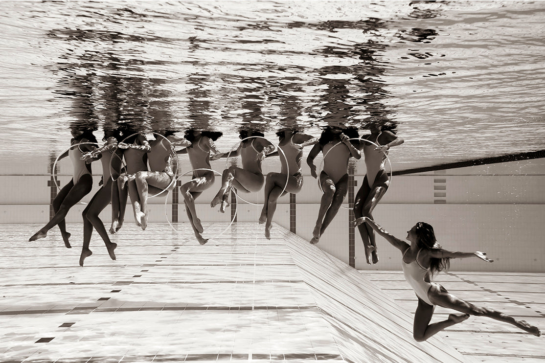 Title: Ladies in Waiting #6
Description: This black and white underwater photo by fine art photographer Francesca Owen captures synchronized swimmers performing. Arranged in a horizontal line, the group executes a dance move using hoops, while one swimmer extends away from the group with arms outstretched. This limited edition piece is printed on Giclee fine art paper.