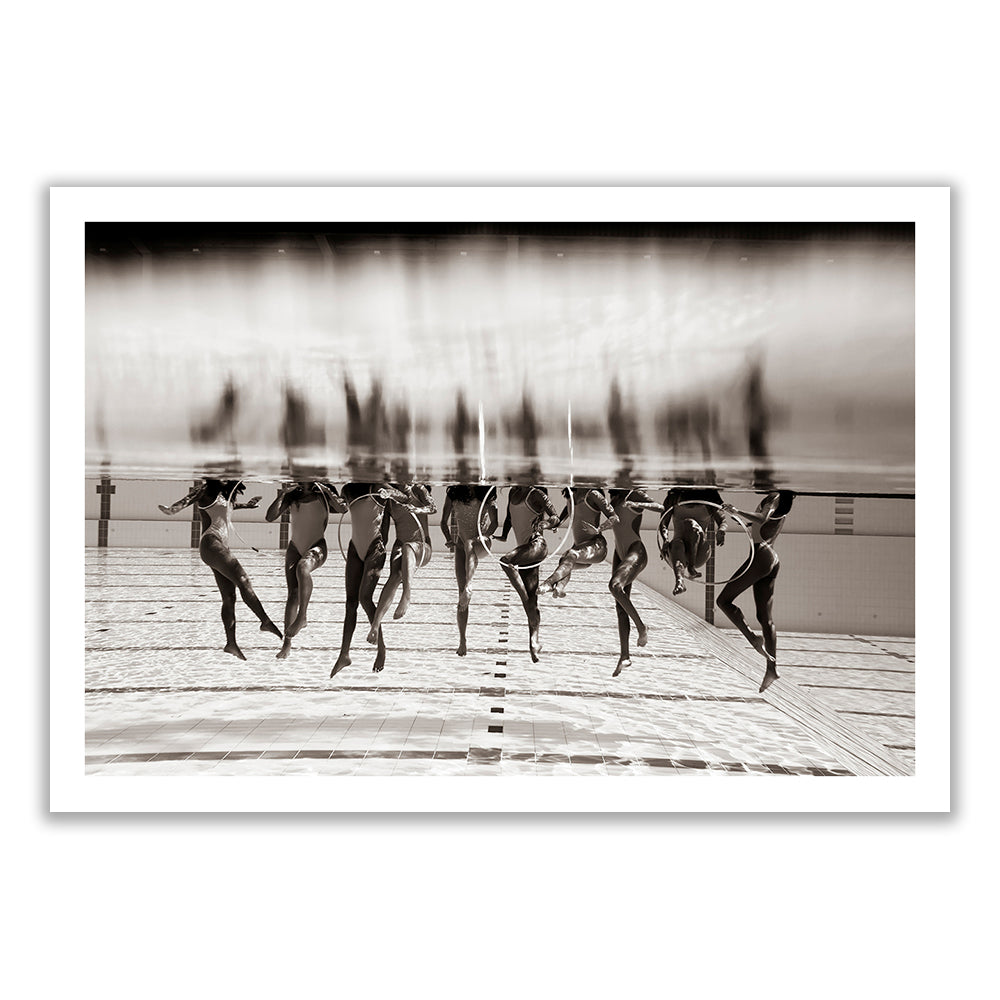 ‘Ladies in Waiting #3’ by Francesca Owen captures a black and white underwater photo of synchronized swimmers performing a routine. The swimmers are elegantly submerged from the hips down, wearing swimsuits and caps, with their legs extended in various formations. This limited edition piece is printed on giclee fine art paper.