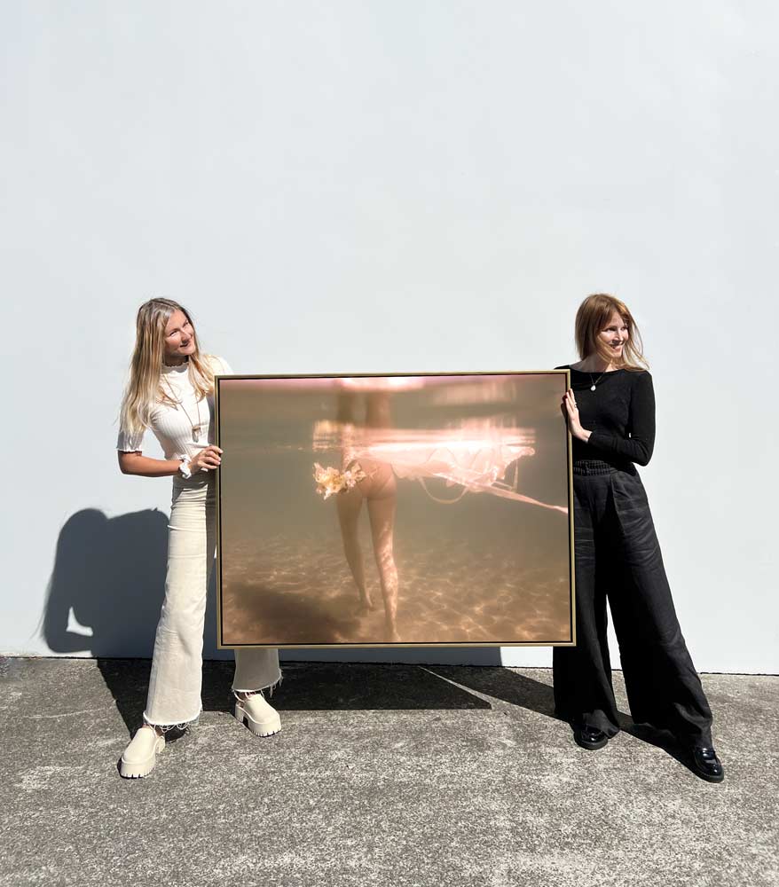 Two women stand against a plain white wall, each holding one side of a large framed photograph. The image within the frame, titled "The Language of Flowers #2" by Francesca Owen, appears to be an abstract or blurred figure with warm tones, possibly underwater. This fine art print is created using archival pigments and casts a shadow on the ground.