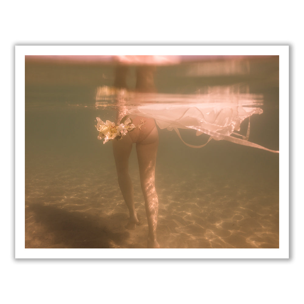 A person submerged underwater with their legs enveloped in the soft folds of a wedding dress, which floats ethereally around them. The image is illuminated by a gentle, warm light, with a bouquet of flowers gracefully drifting near the dress. The scene exudes a serene and dreamy quality, making it an ideal subject for fine art prints produced using archival pigments. This artwork is titled "The Language of Flowers #2" by Francesca Owen.