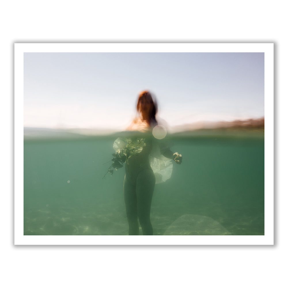 A blurred figure of a person stands waist-deep in water, holding a bouquet of flowers. Captured from an underwater perspective with soft lighting and a clear sky, this image is reminiscent of "The Language of Flowers #3" by Francesca Owen, featuring fine art paper prints enhanced by archival pigments for lasting vibrancy.