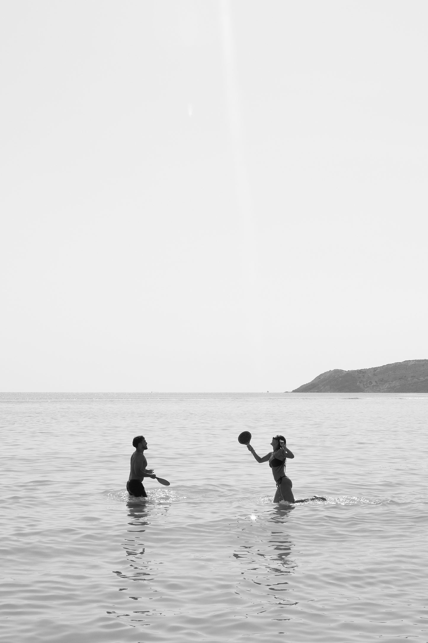 Title: "Magic in Milos #8" by Francesca Owen
Two people are engaging in a playful moment with a beach ball in shallow ocean waters, depicted in striking black and white. One is poised to catch or throw the ball, while the horizon and a distant hill frame the scene. This fine art print uses archival pigments on quality paper and includes a certificate of authenticity.