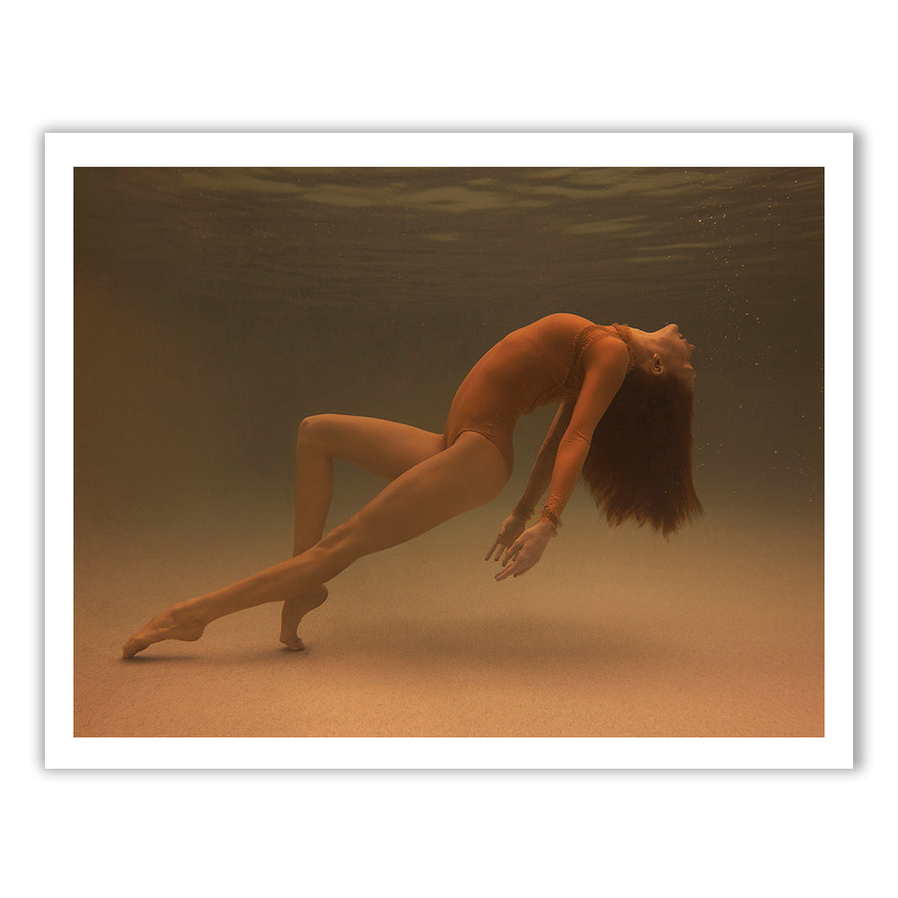 Beneath the water's surface, a figure from Francesca Owen's Open Hearted collection elegantly arches their back in an orange bodysuit. With their head tilted upwards, legs extended, and toes pointed, they create a dynamic and graceful pose against a softly illuminated background. Bubbles trail from their lips as they move.