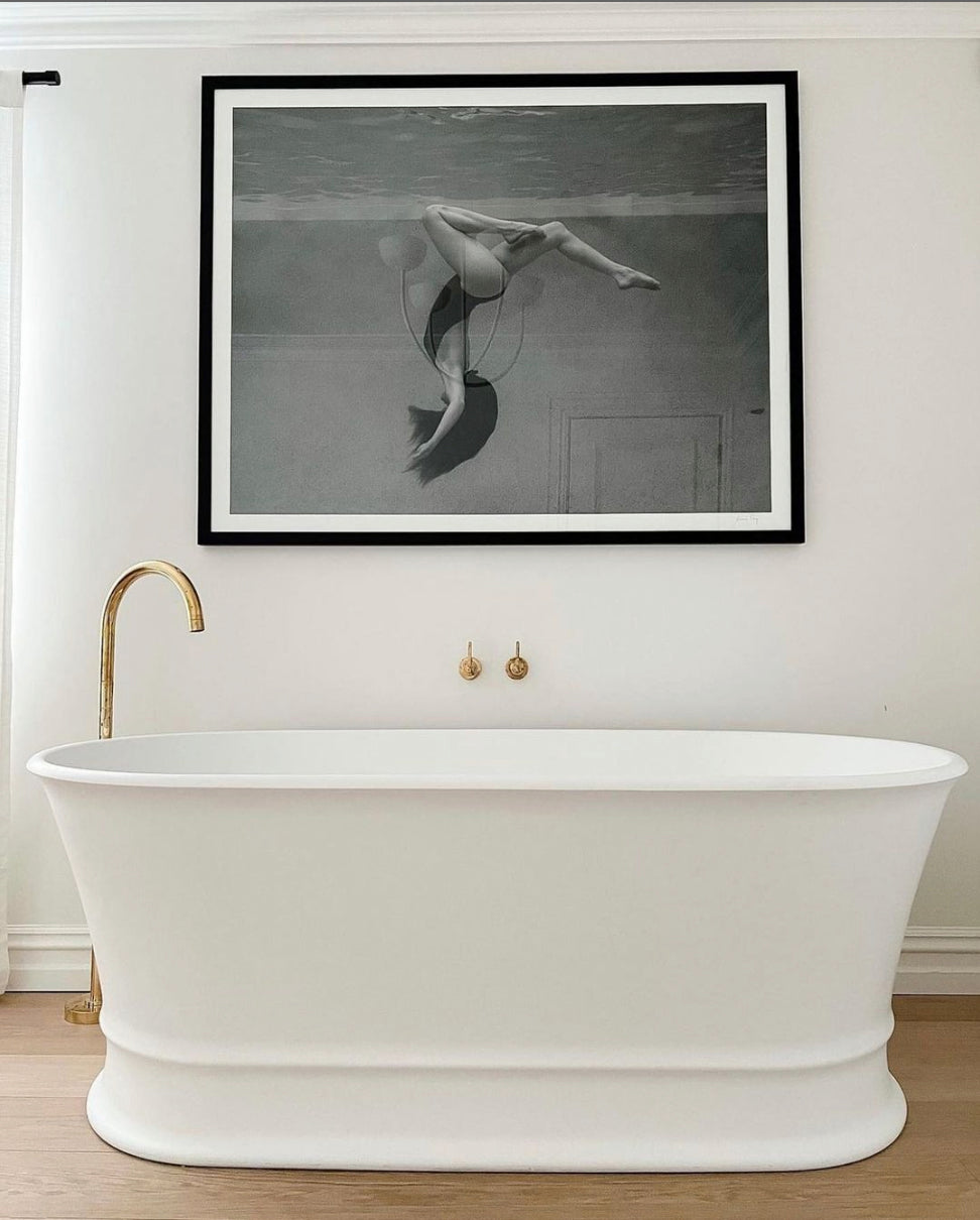 A modern bathroom featuring a sleek white freestanding bathtub, elegant gold fixtures, and Francesca Owen's "Palm Springs Solo" photograph of an underwater swimmer on the wall above the tub. The fine art paper print, created with archival pigments, enhances the minimalist design and is perfectly complemented by the room’s wooden floor.