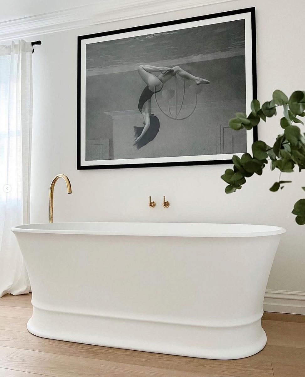 The Palm Springs Solo by Francesca Owen is positioned against a wall below a large black-and-white photograph of a swimmer underwater, printed on fine art paper using archival pigments. This bathroom setup features a gold faucet and handles, a light wooden floor, and a plant with green leaves to the right side.