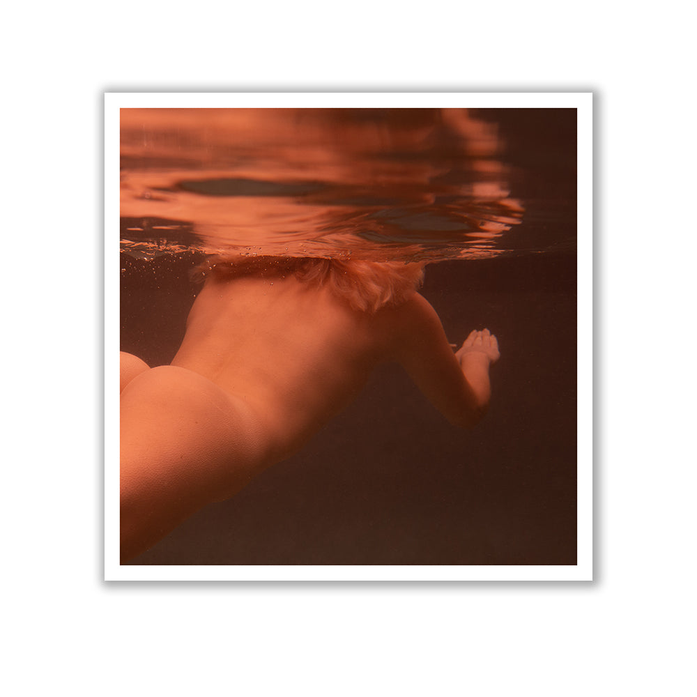 A person with blonde hair is swimming underwater, partially submerged. The water and their skin are illuminated by a warm, orange light. Only the back and partial side of the person's body are visible, as if captured in "Reflect no.3" by Francesca Owen using archival pigments on fine art paper, complete with a certificate of authenticity.