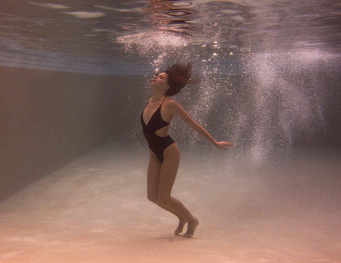 A woman in a black one-piece swimsuit, titled "Self Reflection 03" by Francesca Owen, floats gracefully underwater in a swimming pool, surrounded by bubbles. She appears to be enjoying the serene environment, with her hair flowing freely and her hands extended lightly by her sides—a moment captured perfectly to be printed on fine art paper.