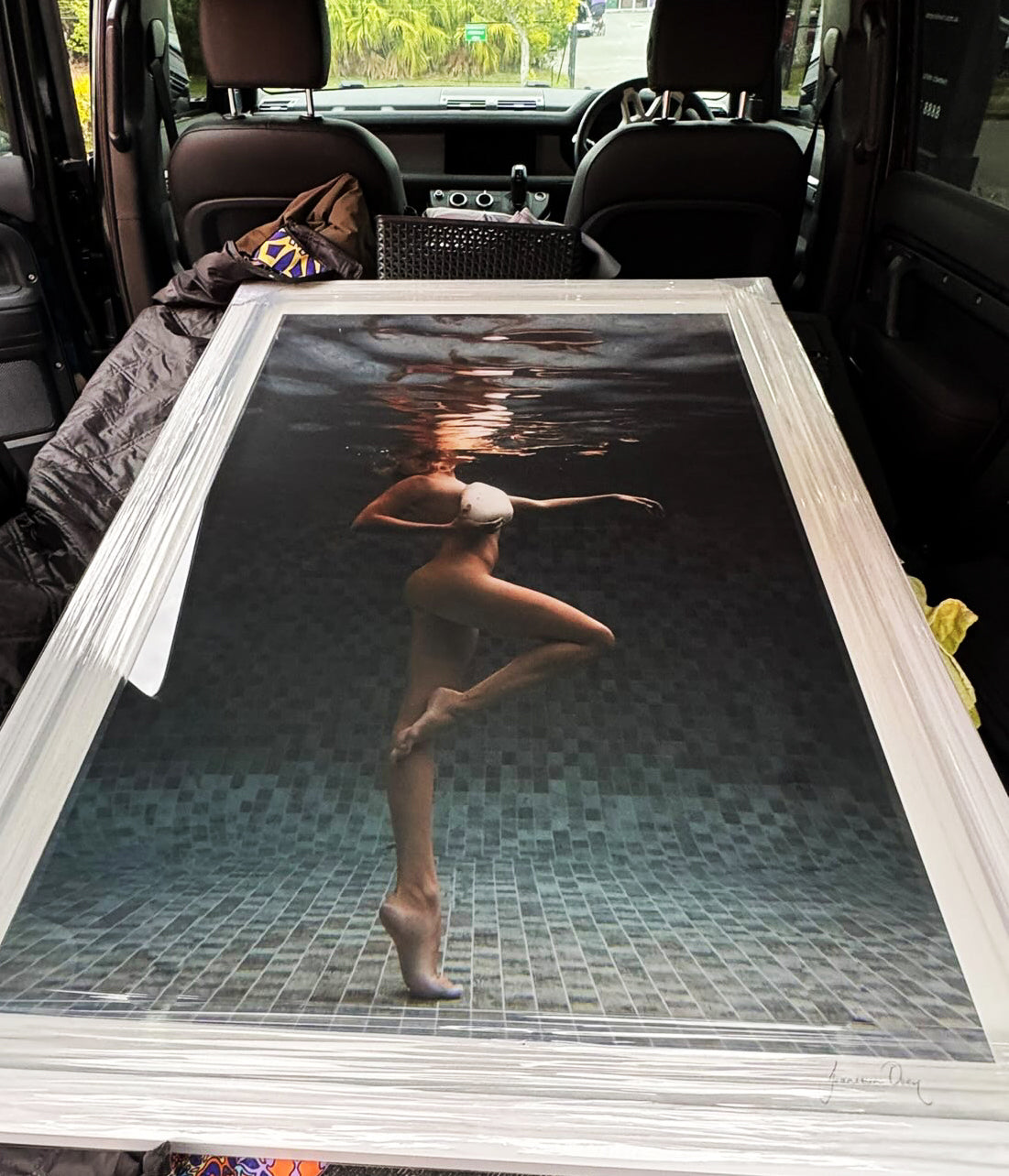 Shell & She" by Francesca Owen is a framed fine art print of a woman underwater in a white bathing cap and swimsuit, posing gracefully in a tiled pool. Includes certificate of authenticity; shown placed in the back of a car.