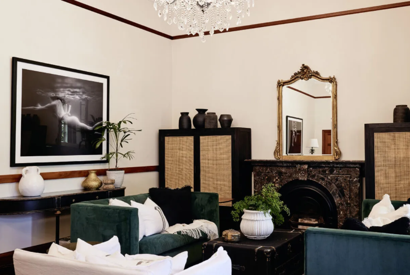 A sophisticated living room features emerald green velvet chairs, a black marble fireplace with a gilded mirror above, and "Gliding #1," an elegant black-and-white photograph by Francesca Owen, printed with archival pigments. The room is adorned with black vases, green plants, and a crystal chandelier.
