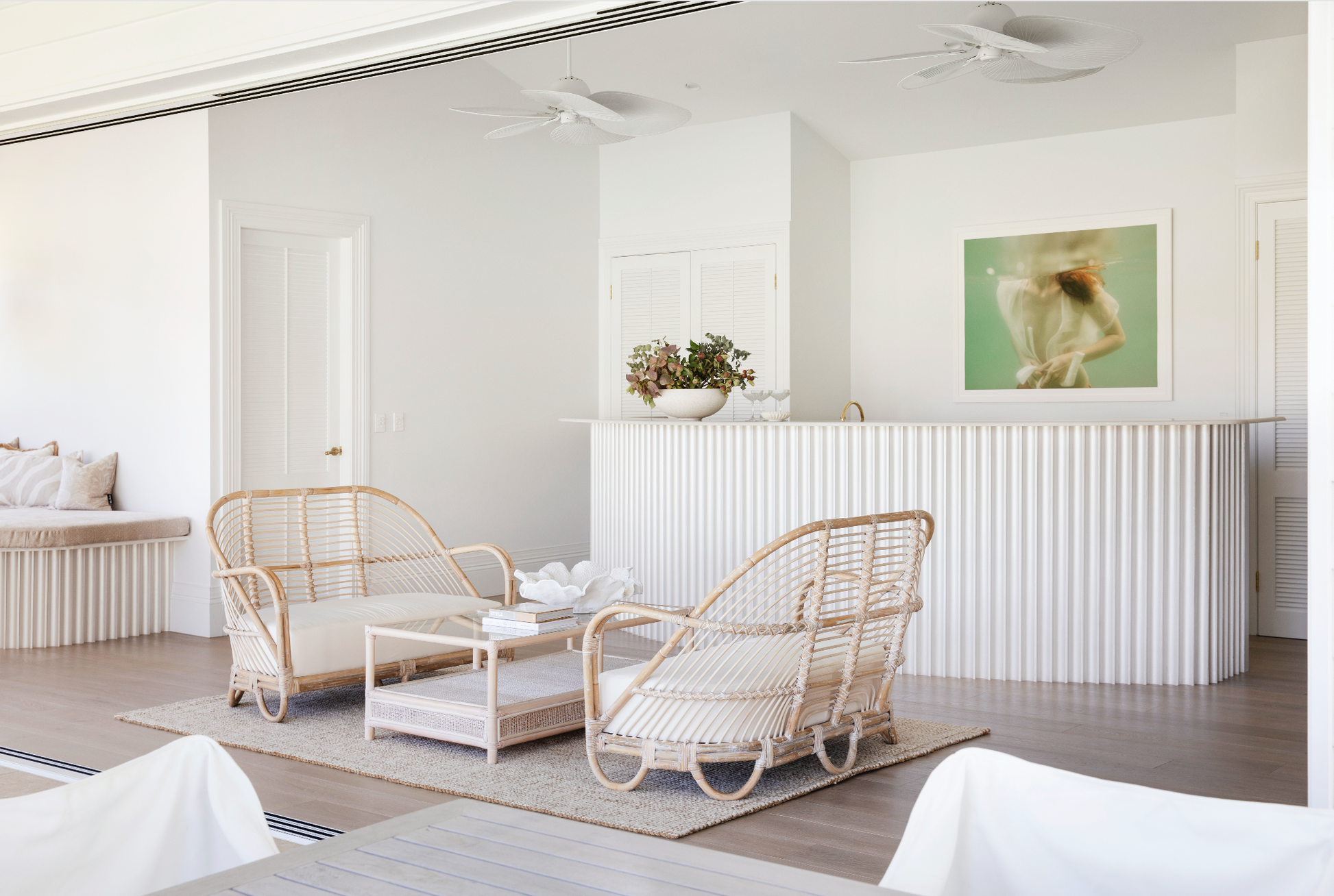 A bright, minimalist living space with a white and wood color scheme. It features wicker armchairs, a small coffee table, and a bar area with potted plants. The room has ceiling fans, a cushioned window seat, and "Softness #1," an underwater-themed photograph by Francesca Owen printed on fine art paper with archival pigments.