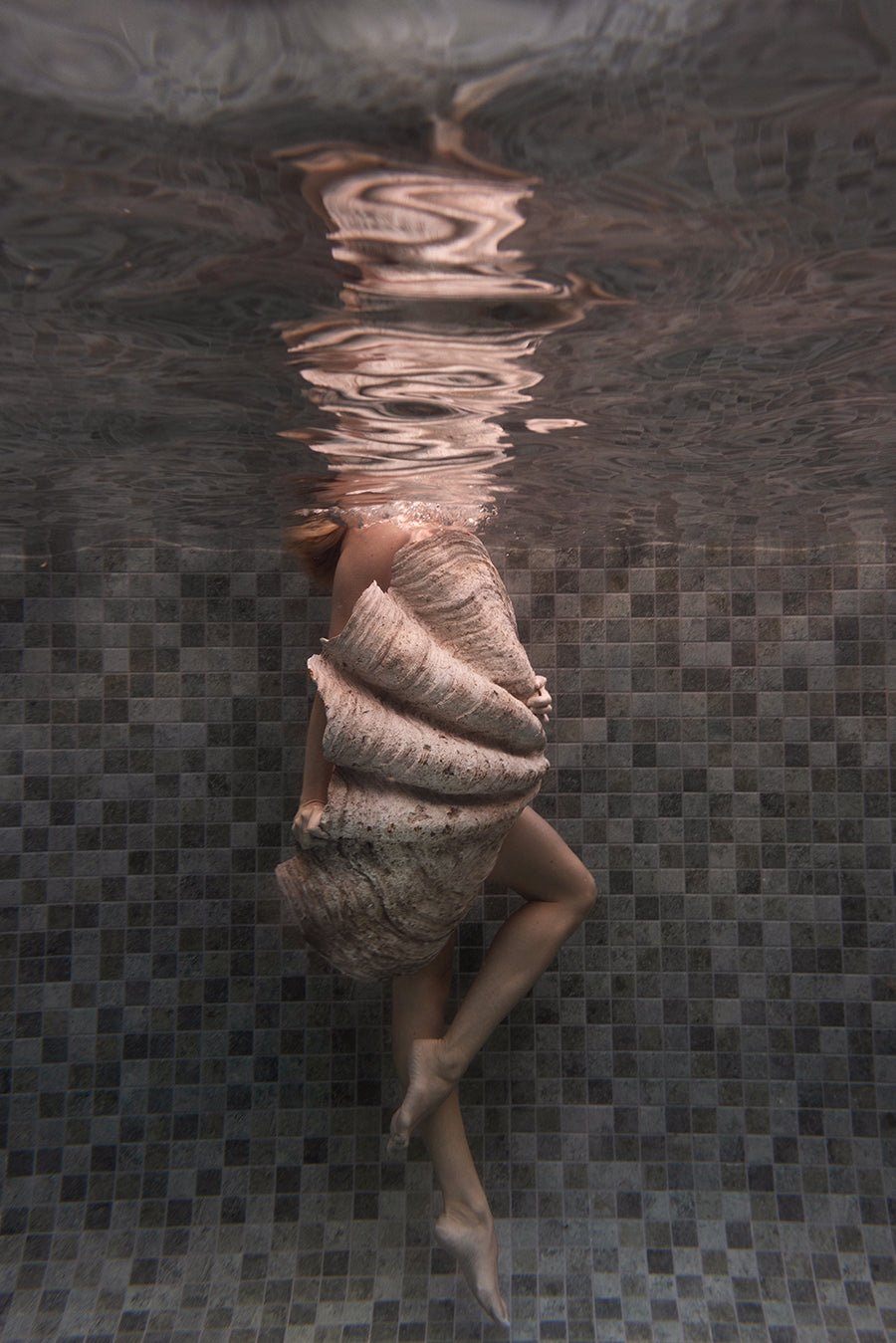 A person in a textured dress floats underwater, their head breaking the surface. Tiled pool walls and rippling reflections create a dreamlike mood in "Shell & She #3"—a fine art print by Francesca Owen with certificate of authenticity.