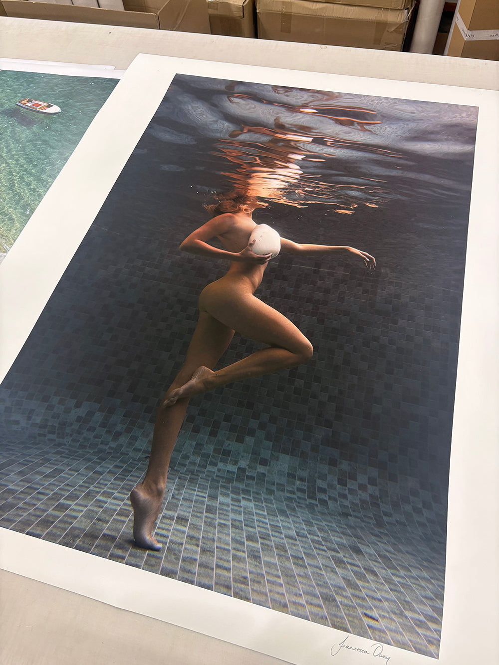 Francesca Owen's "Shell & She" shows a woman underwater in a white bikini, swimming gracefully beneath reflected light. This hand-signed fine art print rests on paper with boxes in the background.