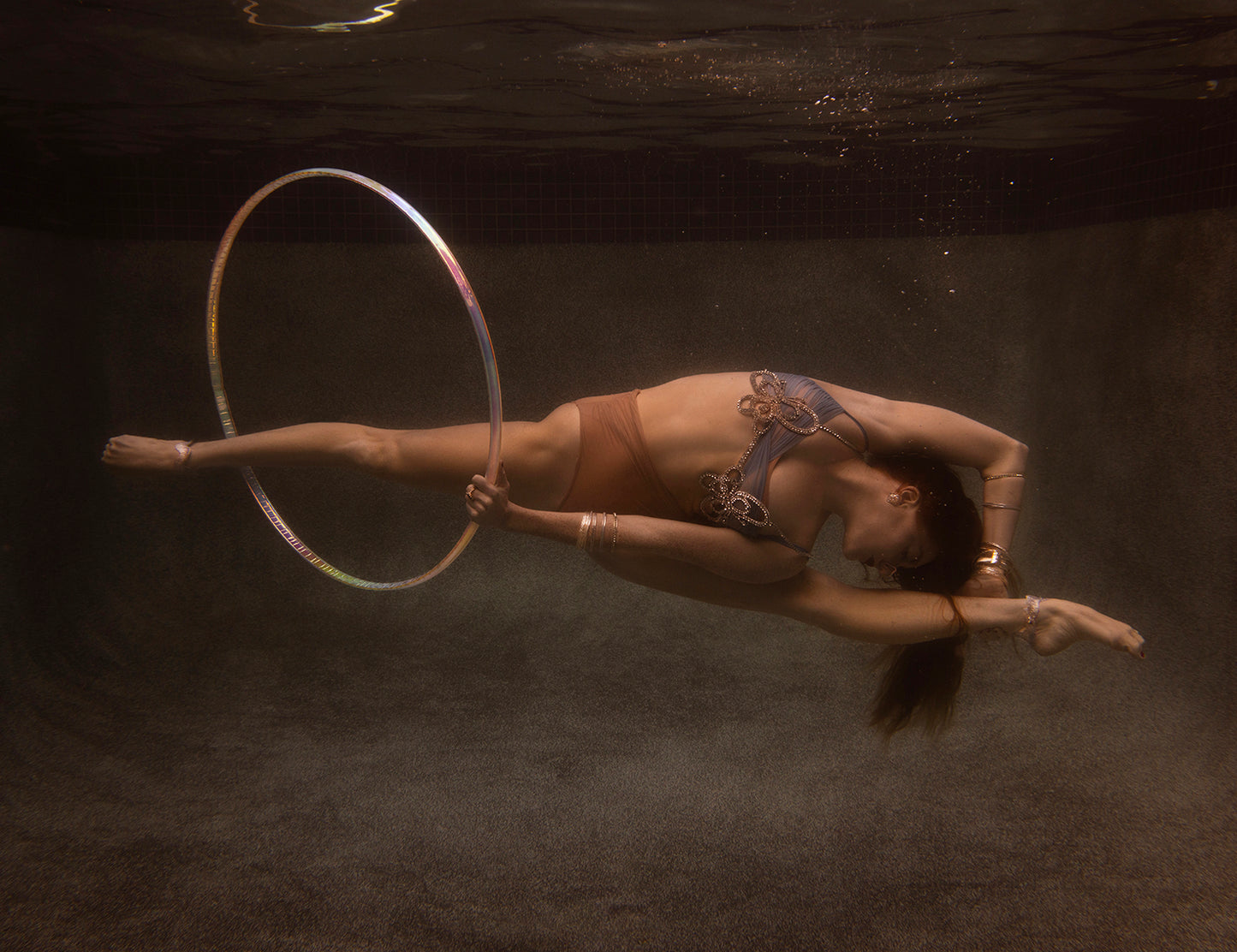 In "Show Time Part 3, #1" by Francesca Owen, a person gracefully balances a hoop on their right leg underwater, arching their back and extending their arms and legs. With its serene and ethereal quality in muted colors reminiscent of fine art prints, custom framing could enhance the artwork's beauty.