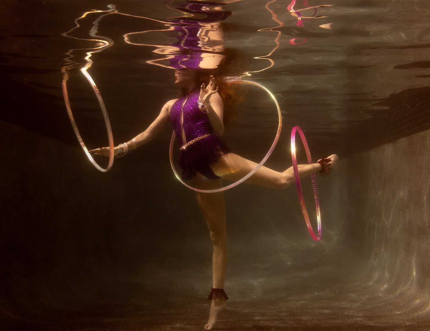 Show Time 2" by Francesca Owen depicts a woman in a purple costume performing underwater with three hula hoops, surrounded by shimmering light—printed with archival pigments on fine art paper.