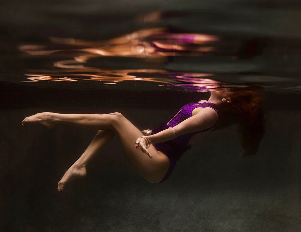 A woman in a purple swimsuit floats underwater with her hair flowing upwards, beautifully reflected on the water's surface. The scene captures the artistry of archival pigments, creating a serene and dreamy atmosphere reminiscent of fine art paper's textured beauty, as seen in "Show Time Part 2, #3" by Francesca Owen.
