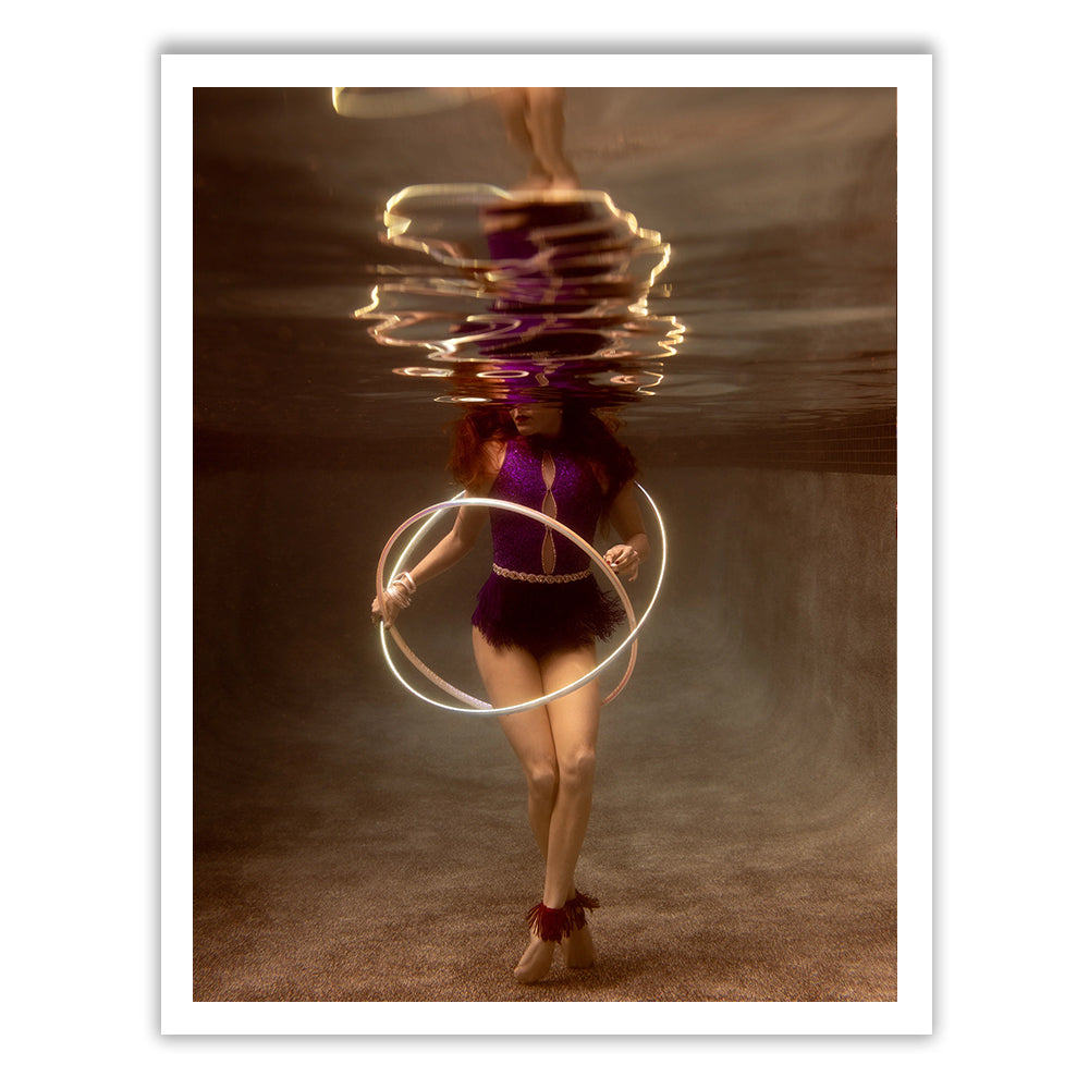 Francesca Owen is submerged in a pool, adorned in an outfit from the Show Time 8 collection, and holding glowing hoops. The water's surface reflects her image, creating a mirrored effect reminiscent of fine art paper using archival pigments and capturing the ethereal scene with timeless elegance.