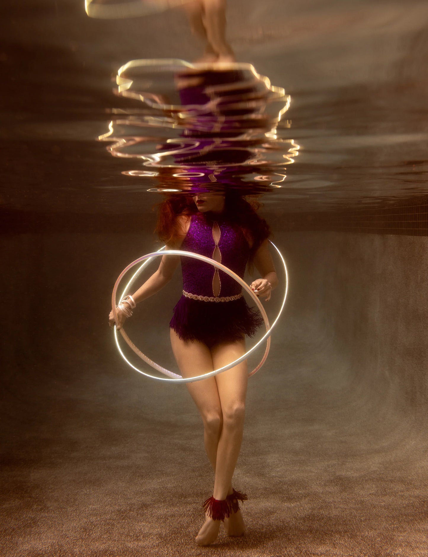 Francesca Owen's Show Time 8 features a captivating underwater scene on fine art paper, depicting a woman in a purple outfit holding an illuminated hula hoop. Her red hair drifts beneath the surface while light reflections cast wavy patterns around her.