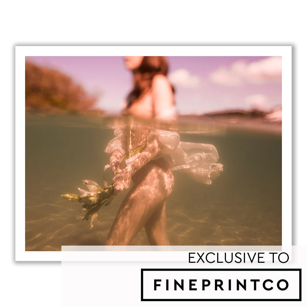 A minimalist, airy living room features a white sofa with brown cushions, a surfboard mounted against the wall, a modern lamp, and a tall plant in a woven basket. Above the sofa hangs "The Language of Flowers #1" by Francesca Owen, framed and printed on fine art paper using archival pigments. A small nook is visible to the left.