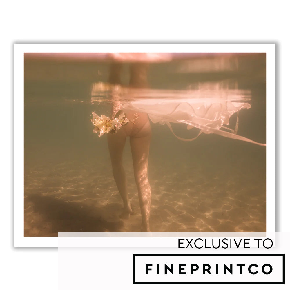 A partially submerged image of a person walking underwater. The water is clear and illuminated with a soft light. The person is holding white flower stems, their legs visible beneath a flowy white dress that floats around them. Text at the bottom reads "EXCLUSIVE TO FINE PRINT CO.", showcasing fine art prints using archival pigments. This exquisite piece is titled "The Language of Flowers #2" by Francesca Owen.