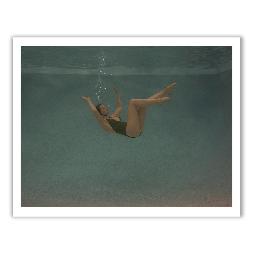 In "Silent Surrender #1" by Francesca Owen, a person in a black swimsuit is floating underwater in a swimming pool, their arms and legs extended gracefully. The background showcases the pool's smooth, light-blue interior. Near the person's mouth, bubbles are visible, captured with archival pigments. The scene exudes calmness and serenity.