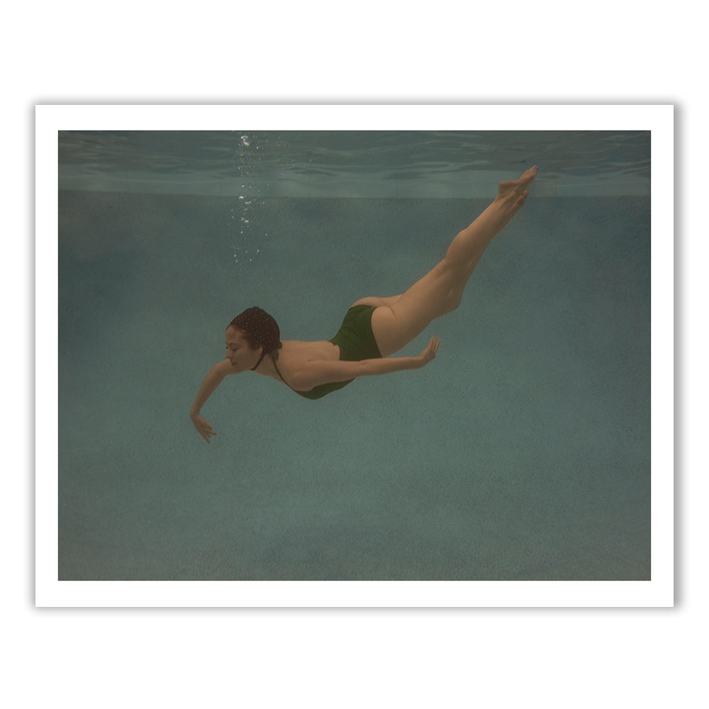 A person in a green swimsuit is swimming underwater in a pool. Their streamlined body and extended limbs create a graceful pose, with bubbles trailing from their mouth as they hold their breath. The pool's blue water provides a serene backdrop, reminiscent of Francesca Owen's fine art print "Silent Surrender #2.